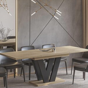 Könnte beinhalten: Ein modernes Esszimmer-Set mit einem hellen Holztisch mit schwarzem V-förmigen Fuß, umgeben von sechs dunkelgrauen Lederstühlen. Der Raum hat eine neutrale Farbpalette mit grauen Wänden und beigen Vorhängen sowie dekorativer Beleuchtung.