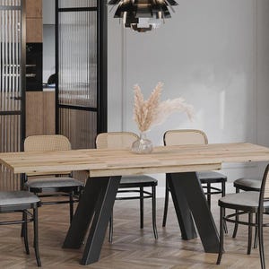 Könnte beinhalten: Ein Esstisch aus hellem Holz mit schwarzen Metallbeinen, umgeben von sechs Stühlen mit gewebten Rückenlehnen und grauen Kissen. Eine Glasvase mit getrockneten Blumen steht auf dem Tisch. Eine moderne Deckenleuchte hängt darüber.