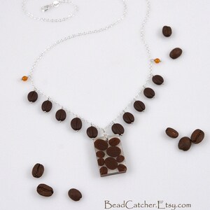 May include: A silver chain necklace with a clear pendant filled with brown coffee beans. The pendant is rectangular and hangs from the chain. There are also brown coffee beans scattered around the necklace.