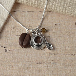 May include: A silver chain necklace with a charm of a small coffee cup and saucer, a coffee bean, and a small spoon. The necklace is on a burlap background with coffee beans scattered around it.