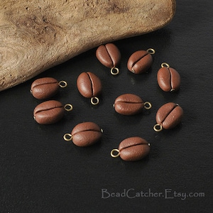May include: A collection of 10 brown coffee bean charms with gold jump rings. The charms are scattered on a black surface.