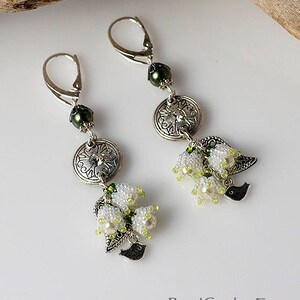 May include: Silver-tone dangle earrings with white beaded flower clusters, green accents, and a small black bird charm. The earrings have a lever-back closure and are displayed on a white surface with a wooden branch in the background. The text "BeadCatcher.Etsy.com" is visible.