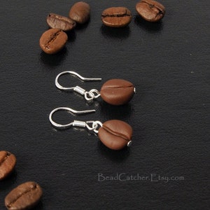 May include: A pair of silver dangle earrings with brown coffee bean charms. The earrings are on a black background with several coffee beans scattered around them.