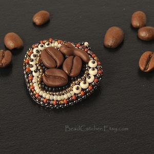 May include: A beaded coffee bean brooch with brown, white, and gold beads. The brooch is shaped like a heart and features a cluster of coffee beans in the center.