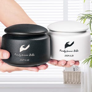 Puede incluir: Dos urnas de cerámica con tapas, una negra y otra blanca. Ambas urnas tienen un diseño de huella de mano y el texto "Family forever Bella 2024.6.26".