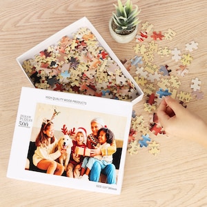 Puede incluir: Un rompecabezas de 500 piezas que presenta a una familia de cuatro con un perro en un ambiente navideño. La caja del rompecabezas es blanca con una imagen del rompecabezas en la parte delantera y el texto "JIGSAW PUZZLES 500 PIECE PUZZLE" y "HIGH QUALITY WOOD PRODUCTS".