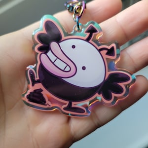 May include: A holographic keychain featuring a cartoon character with a black and white body, pink mouth, and black wings. The character is standing on a small pile of poop.