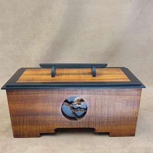 May include: A rectangular wooden box with a dark brown finish and a lighter wood grain top panel. The lid features a black handle. The front side displays a circular cutout containing a dark, textured inlay with a small, light-colored circular accent.