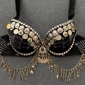 May include: Black bra with silver coin and chain embellishments. The bra features a decorative design with silver coins and green accents. The straps are black and the bra has silver rings on each side.