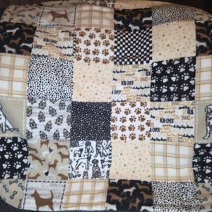 Puede incluir: Una cama para perros de patchwork con varios patrones, incluyendo huellas de patas, siluetas de perros y cuadros escoceses. La paleta de colores incluye beige, marrón, negro y blanco. La cama está diseñada para mascotas y tiene una apariencia acolchada.
