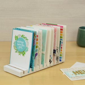 May include: White card holder with a stack of colorful greeting cards. The top card has a blue circle with the text "always here for you" and a floral design.