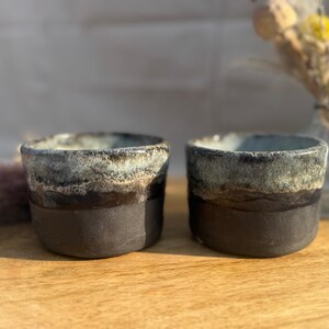 Könnte beinhalten: Zwei handgefertigte Keramikbecher mit dunkelbraunem Boden und einem strukturierten, mehrfarbigen Rand in Blau-, Grün- und Brauntönen. Die Becher stehen auf einer Holzoberfläche, mit einem verschwommenen Hintergrund.
