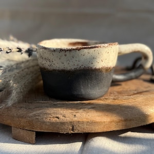 Könnte beinhalten: Eine weiße Keramik-Tasse mit schwarzem Henkel und dem Text "But First, Coffee" in schwarzer, fetter Schrift. Die Tasse steht auf einer weißen Oberfläche.