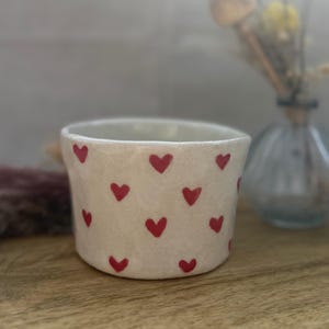 Könnte beinhalten: Ein kleiner, cremefarbener Keramikbecher, verziert mit handgemalten roten Herzen. Der Becher hat eine leicht strukturierte Oberfläche und eine einfache, zylindrische Form. Der Hintergrund ist verschwommen, mit einer Vase mit Trockenblumen.
