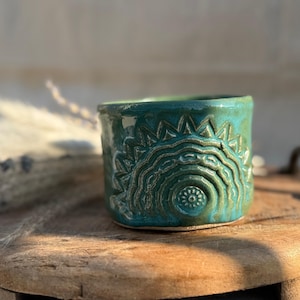 Könnte beinhalten: Handgefertigte türkisfarbene Keramiktasse mit Sonnenmuster. Die Tasse hat eine glänzende Oberfläche und misst etwa 7.6 cm im Durchmesser und 6.3 cm in der Höhe. Das Sonnenmuster ist in die Oberfläche der Tasse geschnitzt.