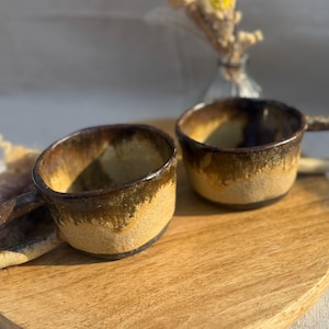 Könnte beinhalten: Zwei handgefertigte Keramikbecher mit einer einzigartigen Glasur. Die Becher haben ein braunes und beiges Farbschema mit einer strukturierten Oberfläche. Sie stehen auf einem Holzservierbrett, was auf ein rustikales oder handwerkliches Thema hindeutet.