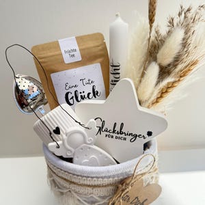 Könnte beinhalten: Ein Geschenk-Korb mit einem weißen Keramikstern und -engel, einer Kerze, einem Teebeutel und Trockenblumen. Auf dem Stern steht "Glücksbringer für Dich". Der Korb ist weiß mit Makramee-Besatz und einem herzförmigen Anhänger.