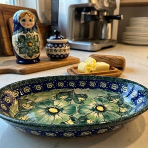 Puede incluir: Una fuente ovalada de cerámica con un diseño floral en tonos azules y verdes. El borde presenta un patrón decorativo. Otros artículos incluyen una tabla de cortar de madera, una muñeca rusa y un tarro pequeño.