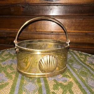 May include: A vintage brass planter with a handle. The oval-shaped planter features a seashell design on the front and decorative floral patterns. The brass has a tarnished patina, giving it a rustic appearance. The handle is arched and attached to the sides.