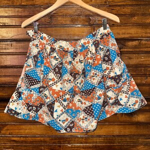 May include: A vintage patchwork skirt with a vibrant mix of colors, including blue, orange, brown, and white. The skirt is hanging on a wooden hanger against a wooden background. The fabric features a repeating pattern of geometric shapes and floral motifs.