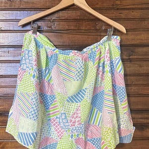 May include: A patchwork skirt in a variety of pastel colors, including pink, blue, and green, hangs from a wooden hanger. The skirt features a mix of geometric patterns, such as stripes, polka dots, and floral designs, creating a vibrant and playful aesthetic.