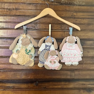May include: Three plush bunny ornaments with fabric dresses hang from a wooden hanger. The bunnies have long ears and are dressed in yellow, blue, and pink dresses with lace and floral details. The background is a wooden wall.