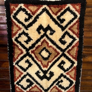May include: A rectangular, hand-tufted rug with a geometric design. The rug features a cream-colored central field with black, brown, and red geometric shapes. The border is black, and the rug is hanging on a wooden hanger.