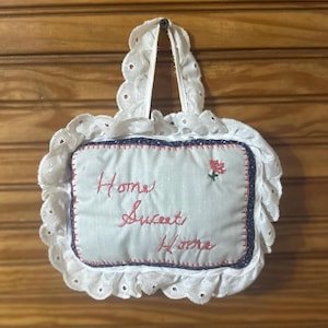 May include: White fabric pillow with a lace trim and embroidered with the text "Home Sweet Home" in red thread. A small red flower is embroidered in the top right corner.