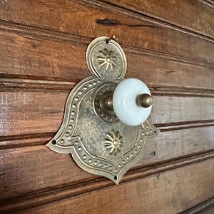 May include: Antique brass door handle with a white porcelain knob. The ornate backplate features a floral design and a decorative border of small circles. The handle is mounted on a wooden door with horizontal panels.