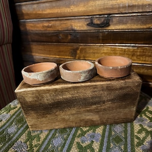 May include: Three small, round, terracotta pots with a weathered appearance sit atop a stained wooden box. The pots have a light-colored rim and a reddish-brown interior. The box is placed on a patterned green and blue fabric.