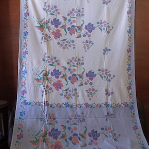 Op de afbeelding: Witte stof met een blauw en oranje bloemenpatroon. De stof heeft een lichtblauwe rand met een bloemenpatroon.