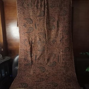 May include: A brown fabric with a paisley pattern hangs on a wooden rod. The fabric has a subtle sheen and a delicate texture.
