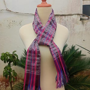 May include: A colourful scarf with a woven pattern in shades of pink, purple, and blue, displayed on a mannequin. The scarf features a detailed design and fringed ends, adding texture. This accessory is suitable for adding a pop of colour to any outfit.