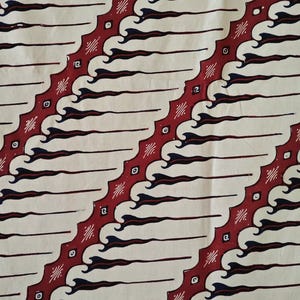 May include: A textile fabric featuring a repeating diagonal pattern of dark red bands with serrated edges. Between these bands are cream-coloured sections decorated with dark blue, flame-like shapes and small, centred square motifs.