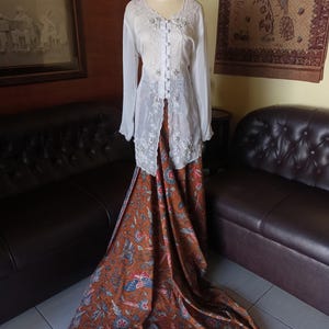 Op de afbeelding: Een witte blouse met lange mouwen, bloemenborduurwerk en knoopsluitingen, gecombineerd met een lange rok met een bruin en blauw bloemenpatroon. Het traditionele kledingstuk wordt op een mannequin getoond.