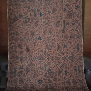 Op de afbeelding: Een bruine stof met een herhalend patroon van blauwe en witte geometrische vormen. De stof heeft een subtiele glans en een zachte textuur.