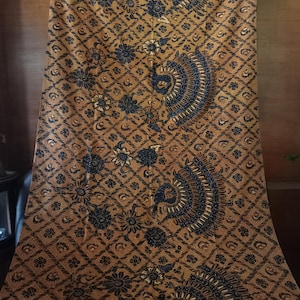 Op de afbeelding: Een bruine en zwarte stof met een herhalend patroon van bloemen en pauwen. De stof is gemaakt van een zacht, lichtgewicht materiaal en heeft een subtiele glans.