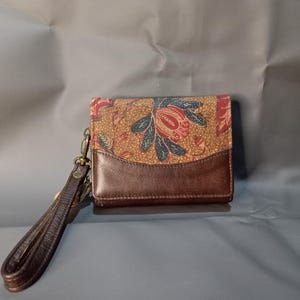 May include: A brown leather wallet with a floral patterned top flap. The pattern includes red, blue, and green accents on a tan background. It has a wrist strap and a brass-coloured clasp.