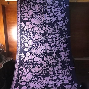 May include: A dark blue textile featuring a light purple floral and bird pattern. The fabric is draped, revealing a detailed design of flowers, leaves, and stylized birds. This textile could be used as a decorative item, such as a curtain or a throw.