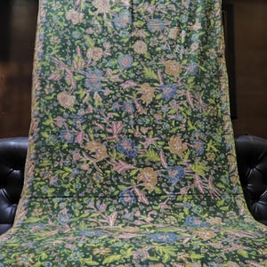 May include: A rectangular fabric textile featuring a dark green base colour with an intricate, repeating floral and botanical pattern in shades of light blue, soft pink, tan, and yellow. The edges are finished with a decorative, patterned border.