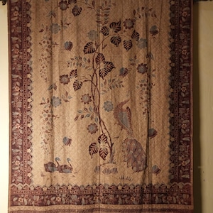 Puede incluir: Textil beige con un detallado estampado floral y de aves. El diseño presenta un árbol central con hojas y flores, y un pavo real y otras aves. Los bordes son de color marrón oscuro con motivos florales. El textil está colgado de una barra de madera.