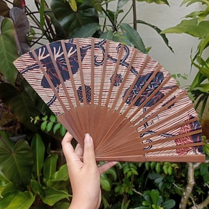 Op de afbeelding: Een bruine, vouwbare houten waaier met een stoffen bekleding met een patroon. De stof is lichtbeige met donkerblauwe en rode bloemenmotieven.