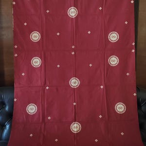Puede incluir: Una pieza de tela rectangular de color granate con un patrón repetitivo de medallones circulares blancos con diseños centrales intrincados. Pequeños acentos blancos en forma de cruz se dispersan por la superficie y un borde decorativo ancho recorre la parte inferior.