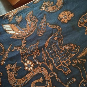 May include: A dark blue fabric featuring a detailed batik pattern in orange and white. The design includes various animals such as a swan, a deer, a tiger, and a serpent, interspersed with floral motifs and leaf shapes across the entire surface.
