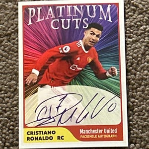 Peut inclure: Carte de collection Platinum Cuts avec Cristiano Ronaldo en maillot de Manchester United. La carte présente une signature fac-similé et le texte "Platinum Cuts" sur un fond coloré.