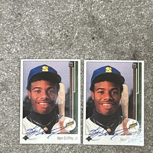May include: Two baseball cards featuring Ken Griffey Jr. The cards show a smiling man wearing a blue baseball cap with a yellow "S" logo. He is holding a baseball bat. The cards have a white border and the text "Ken Griffey Jr."