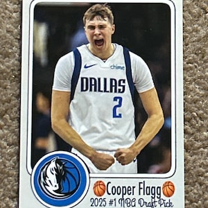 May include: A basketball trading card featuring Cooper Flagg in a white and blue Dallas Mavericks jersey. The card includes the team logo, player's name, and text indicating he is the 2025 #1 NBA Draft Pick. The player is shown with an open mouth.