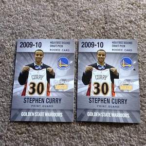 May include: Two Stephen Curry rookie cards from the 2009-10 NBA season. Each card features Curry holding a Golden State Warriors jersey with the number 30. The cards are labeled "NBA First Round Draft Pick Rookie Card" and "Platinum Limited Edition".
