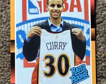 Könnte beinhalten: Eine Steph Curry Rookie-Basketballkarte. Die Karte zeigt Curry, der ein Trikot mit "CURRY" und der Nummer "30" hochhält. Die Karte hat das NBA-Logo und den Text "Rated Rookie". Die Karte ist orange und schwarz umrandet.