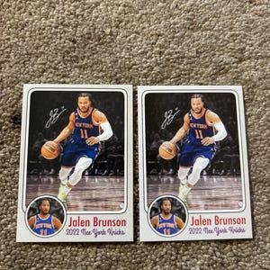 Puede incluir: Dos cromos de Jalen Brunson de los New York Knicks. Cada cromo muestra a Brunson en acción, botando un balón de baloncesto, vistiendo un uniforme azul y naranja. Los cromos incluyen el texto "Jalen Brunson 2022 New York Knicks."